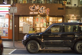 A street view featuring the front of a restaurant with bright signage written in a combination of English and what appears to be Japanese characters. There is a large black SUV parked in front of the restaurant. The restaurant windows display various posters and advertisements, adding to the vibrant and busy atmosphere. Adjacent to the restaurant, a brightly lit storefront with additional posters is visible.