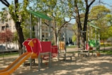 A children's playground featuring a vibrant red and orange slide, a play structure shaped like a bird, and equipment designed for climbing and playing. Surrounding the playground are large trees with green leaves, and several apartment buildings are visible in the background. The area is well-lit with natural sunlight and has a sandy ground covering.