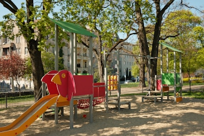 A children's playground featuring a vibrant red and orange slide, a play structure shaped like a bird, and equipment designed for climbing and playing. Surrounding the playground are large trees with green leaves, and several apartment buildings are visible in the background. The area is well-lit with natural sunlight and has a sandy ground covering.