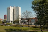 Tall buildings are surrounded by greenery, with a blue sky overhead. The primary focus is a modern hotel building with signage. Trees without leaves occupy the foreground, and other commercial buildings are visible in the background.