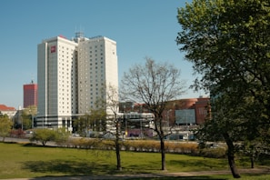 Tall buildings are surrounded by greenery, with a blue sky overhead. The primary focus is a modern hotel building with signage. Trees without leaves occupy the foreground, and other commercial buildings are visible in the background.