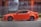 A bright orange sports car is parked next to a metallic fence. The vehicle features customized rims and performance stickers on the windows. Behind the fence, there's a building with large windows and a sign displaying automotive service details.