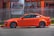 A bright orange sports car is parked next to a metallic fence. The vehicle features customized rims and performance stickers on the windows. Behind the fence, there's a building with large windows and a sign displaying automotive service details.