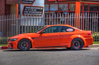 A bright orange sports car is parked next to a metallic fence. The vehicle features customized rims and performance stickers on the windows. Behind the fence, there's a building with large windows and a sign displaying automotive service details.