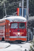 A classic red and orange streetcar is traveling along a track with a rainbow flag in the background. The destination sign on the streetcar reads 'F Market & Castro'. Trees and utility poles surround the area.