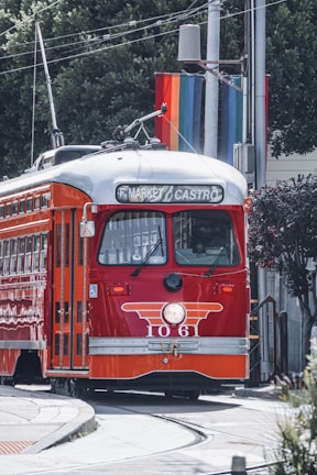 A classic red and orange streetcar is traveling along a track with a rainbow flag in the background. The destination sign on the streetcar reads 'F Market & Castro'. Trees and utility poles surround the area.
