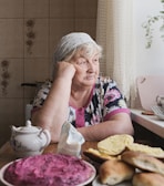 An elderly woman with gray hair and a headscarf sits at a table by a window, gazing thoughtfully outside. The table is set with various foods, including bread rolls and a colorful beet salad. The room has a cozy atmosphere with floral decorations on the walls.