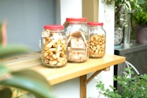 Three glass jars with red lids are placed on a wooden shelf. Each jar contains different types of snacks: pasta in the first jar, what appears to be biscuits or cookies in the second, and small crunchy nuts or crackers in the third. The setting includes surrounding greenery and a rustic outdoor vibe.