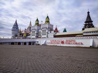 An ornate, colorful complex featuring traditional Russian architectural styles, including towers with vibrant domes and intricate details. The foreground has a wide, paved area leading to a white wall adorned with red Cyrillic lettering.