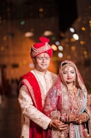 A couple dressed in traditional Indian wedding attire. The groom wears an ornate cream and gold sherwani with a red turban and dupatta, while the bride is adorned in a richly embroidered red and gold lehenga with intricate jewelry including a maang tika and necklace. The warm lighting and blurred background give a festive ambiance.