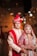 A couple dressed in traditional Indian wedding attire. The groom wears an ornate cream and gold sherwani with a red turban and dupatta, while the bride is adorned in a richly embroidered red and gold lehenga with intricate jewelry including a maang tika and necklace. The warm lighting and blurred background give a festive ambiance.