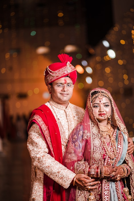 A couple dressed in traditional Indian wedding attire. The groom wears an ornate cream and gold sherwani with a red turban and dupatta, while the bride is adorned in a richly embroidered red and gold lehenga with intricate jewelry including a maang tika and necklace. The warm lighting and blurred background give a festive ambiance.