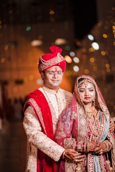 A couple dressed in traditional Indian wedding attire. The groom wears an ornate cream and gold sherwani with a red turban and dupatta, while the bride is adorned in a richly embroidered red and gold lehenga with intricate jewelry including a maang tika and necklace. The warm lighting and blurred background give a festive ambiance.