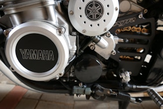 Close-up view of a motorcycle engine showcasing mechanical details, including a prominent Yamaha logo on the left side. The metallic components feature a series of bolts and pipes, with visible parts of a chain and other motorcycle elements.