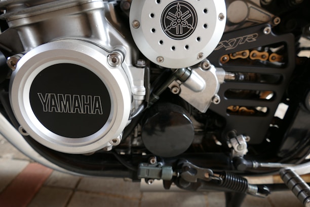 Close-up view of a motorcycle engine showcasing mechanical details, including a prominent Yamaha logo on the left side. The metallic components feature a series of bolts and pipes, with visible parts of a chain and other motorcycle elements.