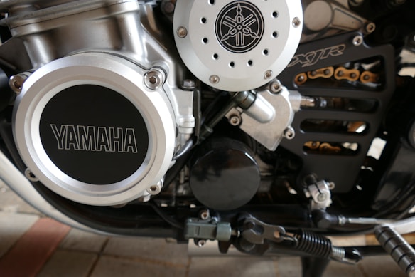 Close-up view of a motorcycle engine showcasing mechanical details, including a prominent Yamaha logo on the left side. The metallic components feature a series of bolts and pipes, with visible parts of a chain and other motorcycle elements.