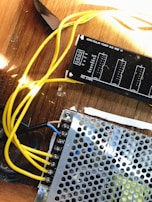 A close-up view of electronic components including a power distribution board labeled 'Moraydular Power Bus Wide V1' and a metal perforated cover. Yellow wires are connected to various terminals, lying on a wooden surface.