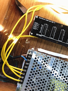 A close-up view of electronic components including a power distribution board labeled 'Moraydular Power Bus Wide V1' and a metal perforated cover. Yellow wires are connected to various terminals, lying on a wooden surface.