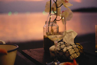 A rustic wooden table set outdoors by the lagoon, laden with fresh seafood dishes and glasses of white wine glowing in the sunset light.