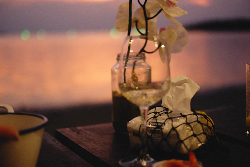 A rustic wooden table set outdoors by the lagoon, laden with fresh seafood dishes and glasses of white wine glowing in the sunset light.