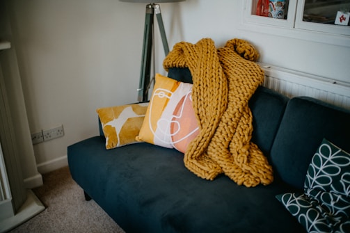 A cozy living room area featuring a dark teal sofa adorned with decorative cushions in bold patterns and colors. A chunky mustard yellow knit throw is casually placed on the sofa. A modern floor lamp stands in the corner, and the room has a warm, inviting atmosphere.