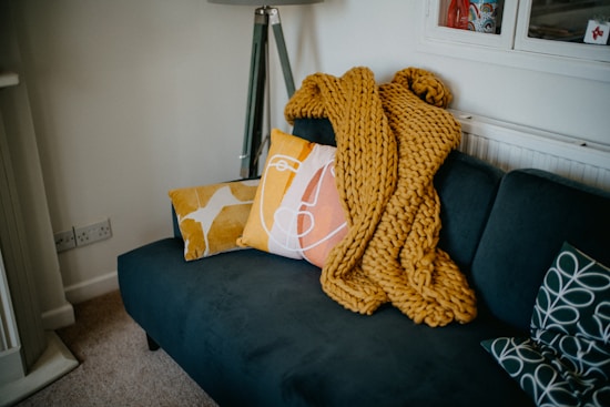 A cozy living room area featuring a dark teal sofa adorned with decorative cushions in bold patterns and colors. A chunky mustard yellow knit throw is casually placed on the sofa. A modern floor lamp stands in the corner, and the room has a warm, inviting atmosphere.