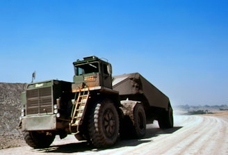 A large, heavy-duty dump truck carries a load of earth or gravel along a dirt road in what appears to be a mining or construction site. The truck's massive tires are dusty, and the sky above is a clear blue, suggesting a bright, sunny day.