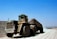 A large, heavy-duty dump truck carries a load of earth or gravel along a dirt road in what appears to be a mining or construction site. The truck's massive tires are dusty, and the sky above is a clear blue, suggesting a bright, sunny day.