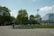 A row of electric scooters is lined up on a paved area near a park, with lush green trees providing a backdrop. In the distance, a modern glass building contrasts with the natural setting. A few pedestrians are visible, enjoying the open space under a clear sky with scattered clouds.