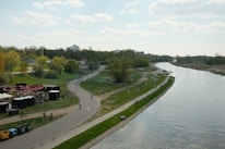 A scenic riverside view with a wide pathway running alongside the river. People are leisurely walking, cycling, and relaxing on the grass. The area is lined with trees, and a small cluster of buildings can be seen in the background. A few booths or tents are set up near the pathway.