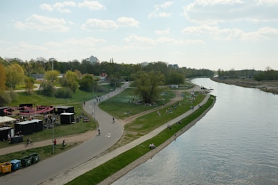 A scenic riverside view with a wide pathway running alongside the river. People are leisurely walking, cycling, and relaxing on the grass. The area is lined with trees, and a small cluster of buildings can be seen in the background. A few booths or tents are set up near the pathway.