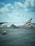 A large commercial airplane from the Emirates fleet is parked on the tarmac under a partly cloudy sky. The aircraft is positioned near the terminal with its engines clearly visible. The tail features the airline's distinctive red, green, black, and white colors. The foreground includes a baggage cart on the left side of the image.