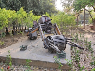 A robotic sculpture made of scrap metal is placed on a concrete platform in an outdoor park setting. The sculpture resembles a futuristic vehicle or robot, featuring cylindrical parts, pipes, and jointed limbs. Surrounding the sculpture are leafy trees and small green shrubs, with fallen leaves scattered on the ground. A green crate is visible near the sculpture.