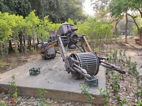 A robotic sculpture made of scrap metal is placed on a concrete platform in an outdoor park setting. The sculpture resembles a futuristic vehicle or robot, featuring cylindrical parts, pipes, and jointed limbs. Surrounding the sculpture are leafy trees and small green shrubs, with fallen leaves scattered on the ground. A green crate is visible near the sculpture.