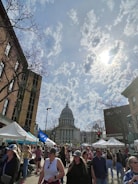 A bustling street market filled with people walking past vendors and white tents. In the background stands a large, domed government building, with bare trees and cloudy skies visible above.