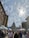 A bustling street market filled with people walking past vendors and white tents. In the background stands a large, domed government building, with bare trees and cloudy skies visible above.