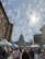 A bustling street market filled with people walking past vendors and white tents. In the background stands a large, domed government building, with bare trees and cloudy skies visible above.