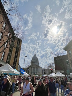 A bustling street market filled with people walking past vendors and white tents. In the background stands a large, domed government building, with bare trees and cloudy skies visible above.