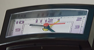 A close-up view of the face of a clock with a non-standard, semi-circular shape. The clock features multi-colored hands in red, yellow, and black, each pointing towards numbers displayed in purple on a light background.