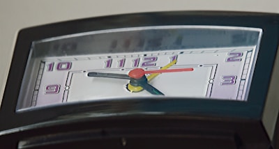 A close-up view of the face of a clock with a non-standard, semi-circular shape. The clock features multi-colored hands in red, yellow, and black, each pointing towards numbers displayed in purple on a light background.
