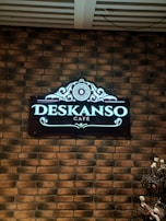 A decorative sign with elegant designs presents the name 'Deskanso Café' in illuminated letters. The sign is mounted on a brick wall, adding to a rustic and cozy ambiance.