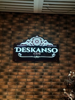 A decorative sign with elegant designs presents the name 'Deskanso Café' in illuminated letters. The sign is mounted on a brick wall, adding to a rustic and cozy ambiance.