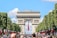 A large, historic monument is flanked by lush green trees lining a busy avenue. The monument is made of stone with detailed carvings and features a prominent archway in the center. Numerous people fill the street, which is decorated with red, white, and blue flags.