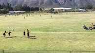 A large open grassy field surrounded by trees and mountains, featuring numerous people engaged in various activities. In the foreground, a group of children are playing with a ball, while others are gathered in groups or walking around in the distance. The scene conveys a sense of community and outdoor recreation.