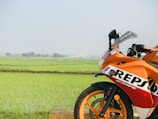 An orange sport motorcycle with REPSOL branding is parked near a lush green paddy field. The motorcycle's front section is visible, showcasing the wheel, handlebar, and part of the body. The background features expansive fields with rows of rice plants extending into the horizon under a clear sky.