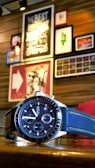 A wristwatch with a black and silver design is placed on a wooden surface. In the background, there is a wall adorned with various framed art pieces and posters, creating an eclectic and cozy atmosphere.