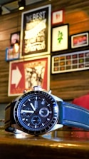 A wristwatch with a black and silver design is placed on a wooden surface. In the background, there is a wall adorned with various framed art pieces and posters, creating an eclectic and cozy atmosphere.