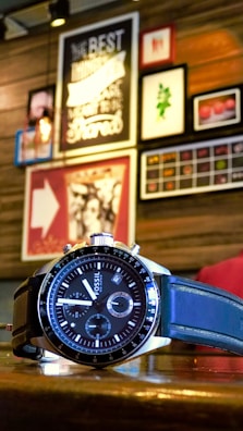 A wristwatch with a black and silver design is placed on a wooden surface. In the background, there is a wall adorned with various framed art pieces and posters, creating an eclectic and cozy atmosphere.