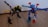 Two colorful robotic figures made from interlocking plastic pieces stand on a wooden floor. One resembles a bird-like dinosaur with a long neck and a beak, while the other resembles a humanoid robot with arms outstretched. They feature a variety of colors including blue, red, yellow, green, and black.