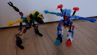 Two colorful robotic figures made from interlocking plastic pieces stand on a wooden floor. One resembles a bird-like dinosaur with a long neck and a beak, while the other resembles a humanoid robot with arms outstretched. They feature a variety of colors including blue, red, yellow, green, and black.